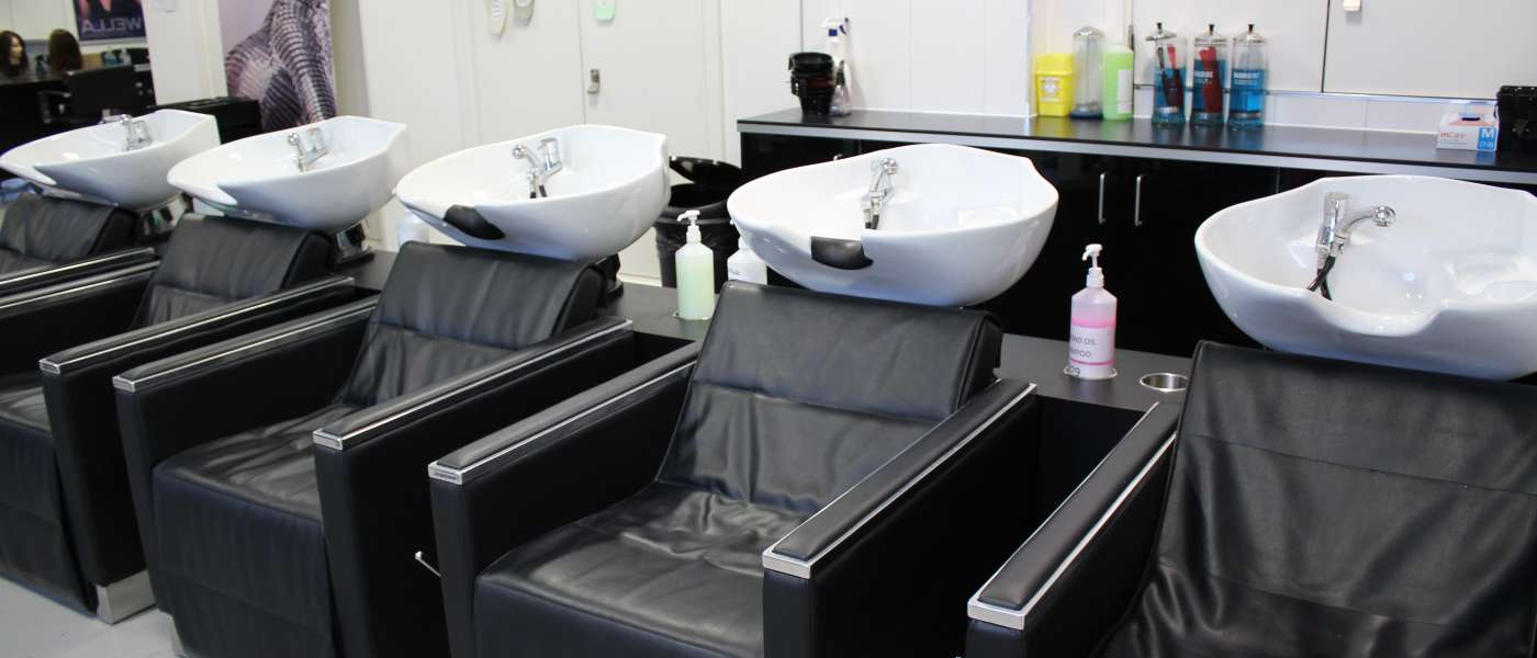 Five black hair salon chairs with attached white sinks. Hair products in the back on a storage unit. Canvas image with a model on the wall and hair mannequins in the far back