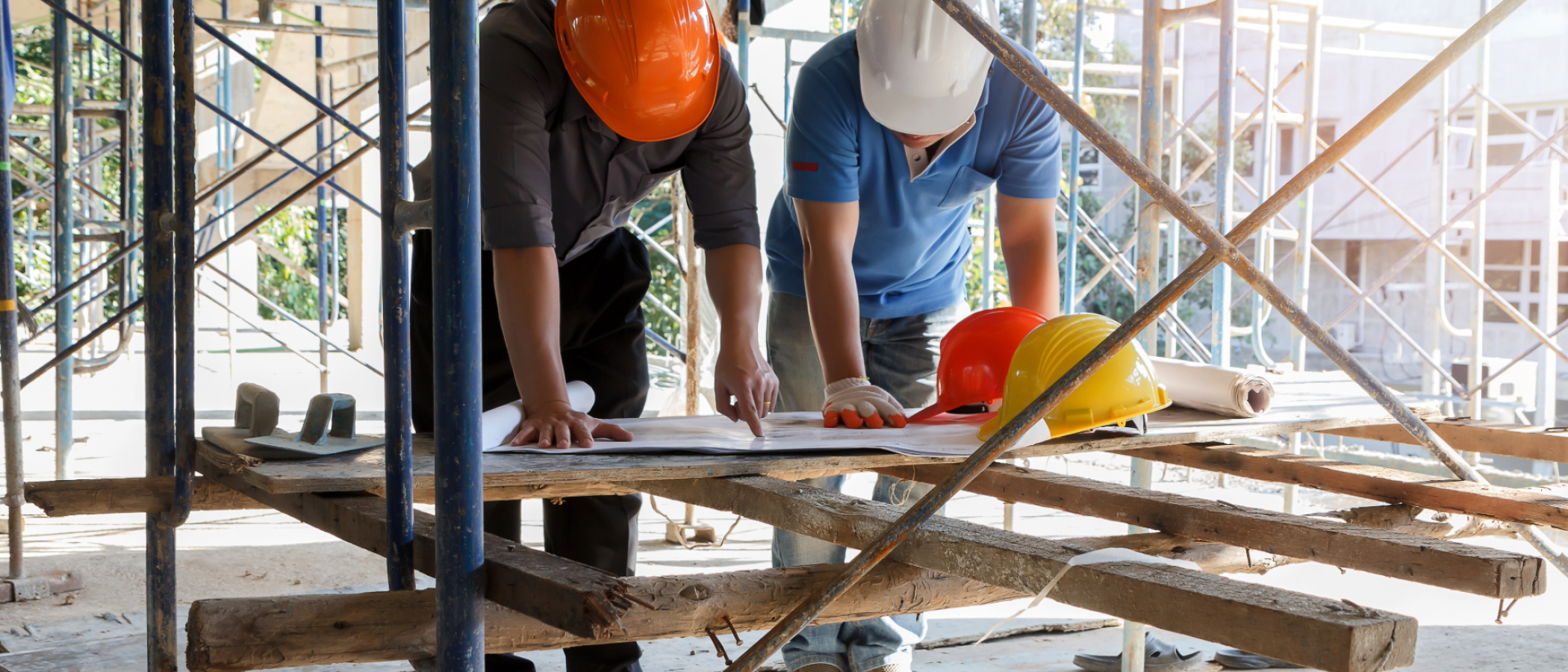 Two construction workers wearing hard hats review building plans on a workbench surrounded by scaffolding at an active construction site.  Provide your feedback on BizChat