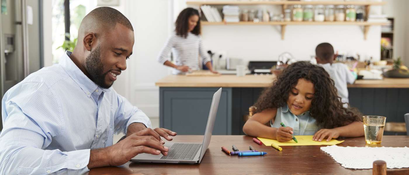 Family in their kitchen man working on laptop at table with girl next to him drawing