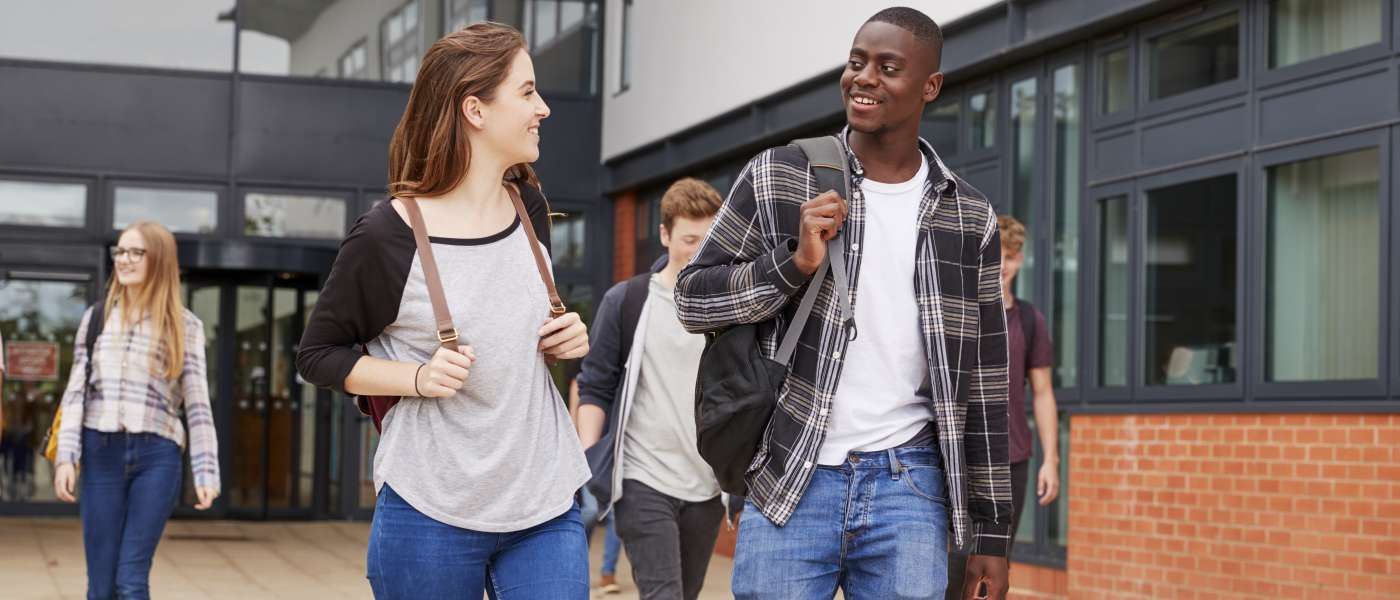 One female student and one male student holding backpacks walking together talking. Three students behind them walking in the same direction outside of a building