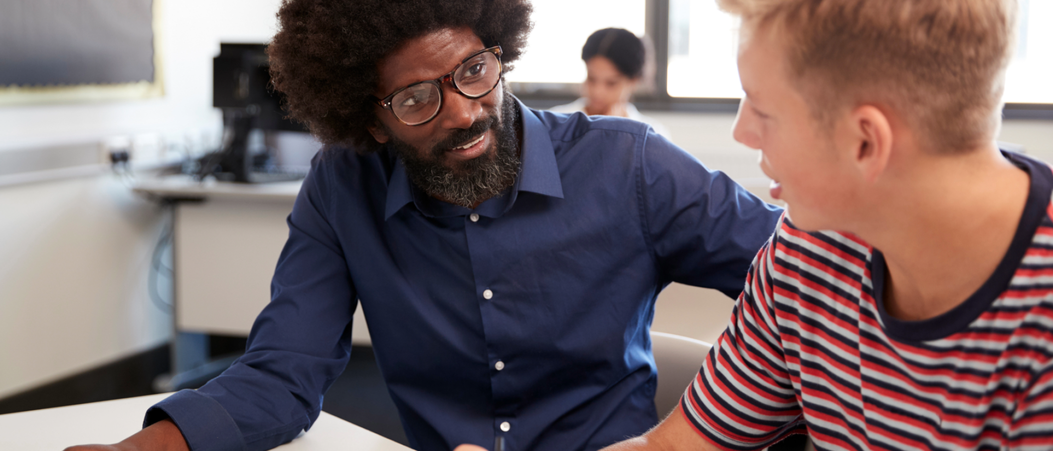 A tutor provides one-to-one guidance to a student at a classroom desk, reviewing written work and offering academic support in a learning environment.