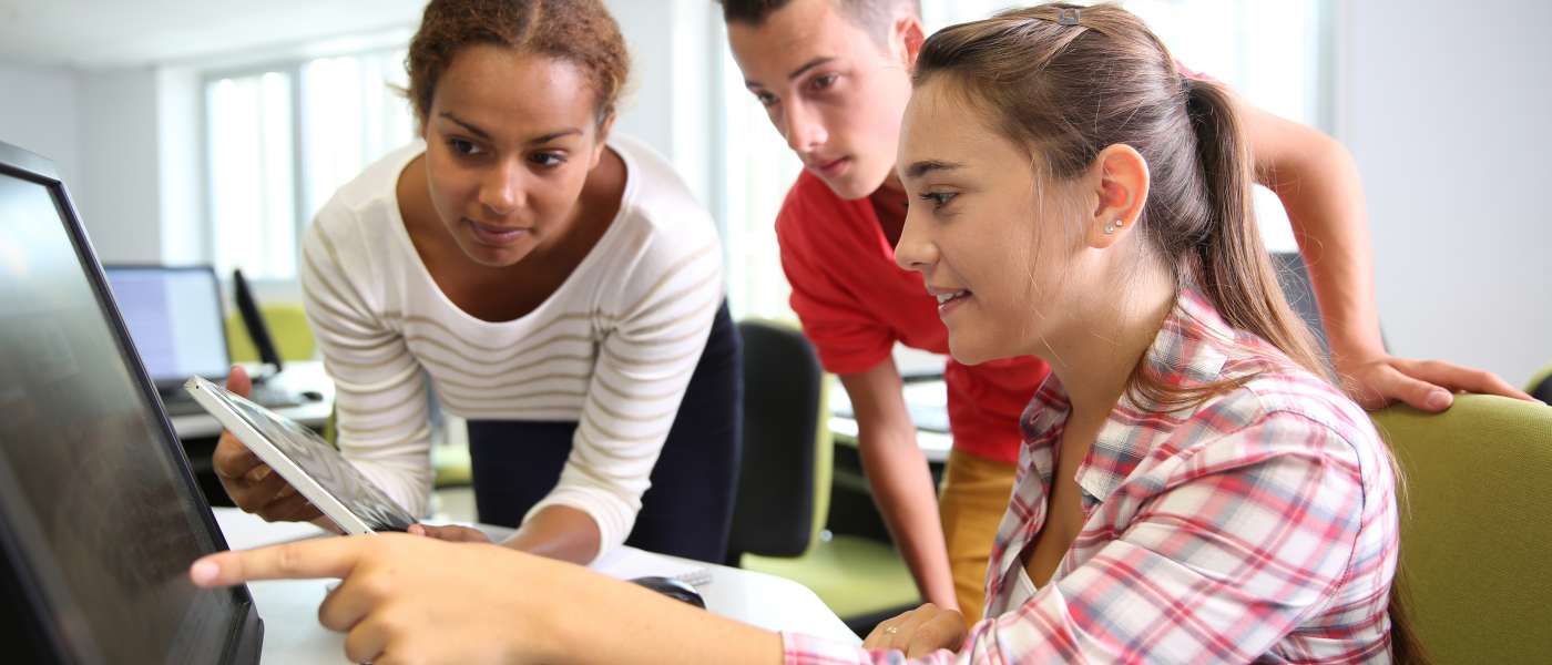 Three learners - one male and two female looking at a computer screen. One female is sat down and pointing at the screen explaining something to the other two learners who are standing up