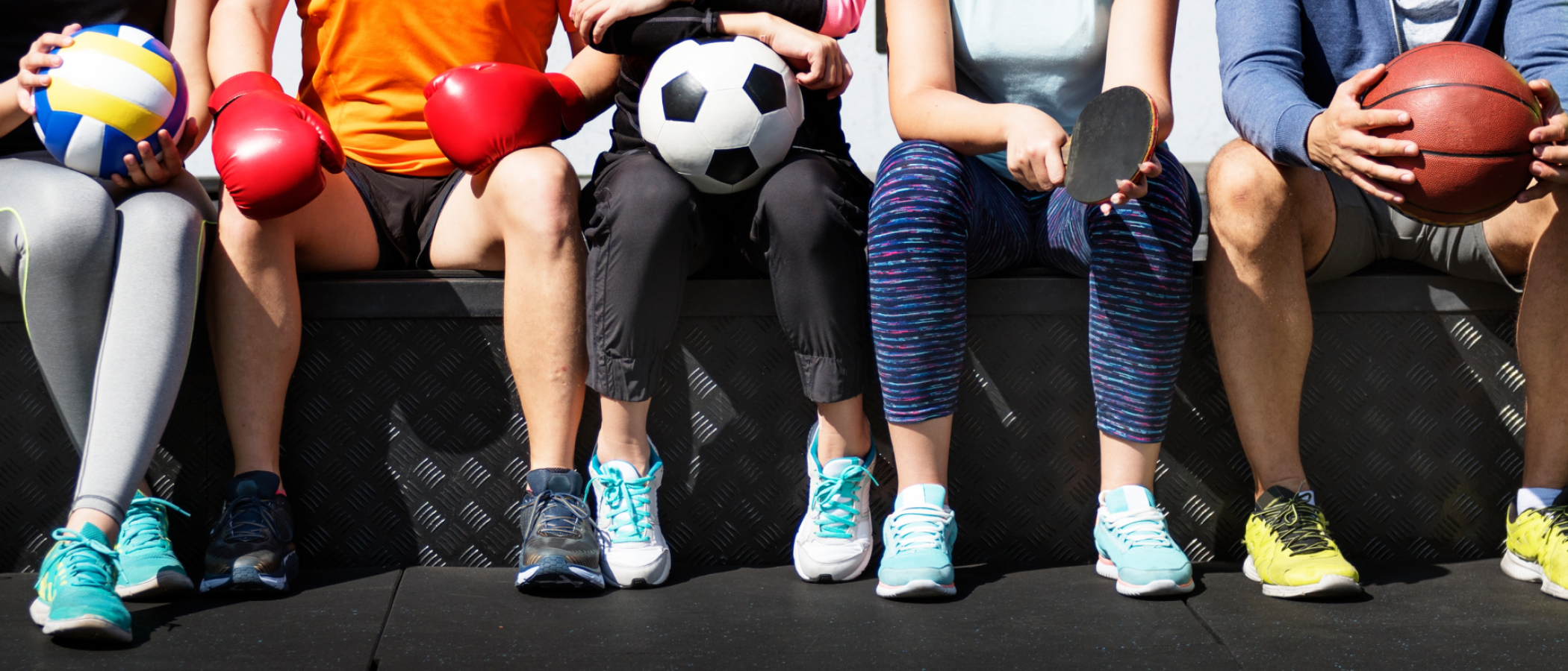 A group of people sit side by side holding different sports equipment, including a volleyball, football, basketball, table tennis bat and boxing gloves, representing participation in a range of sports and physical activities.
