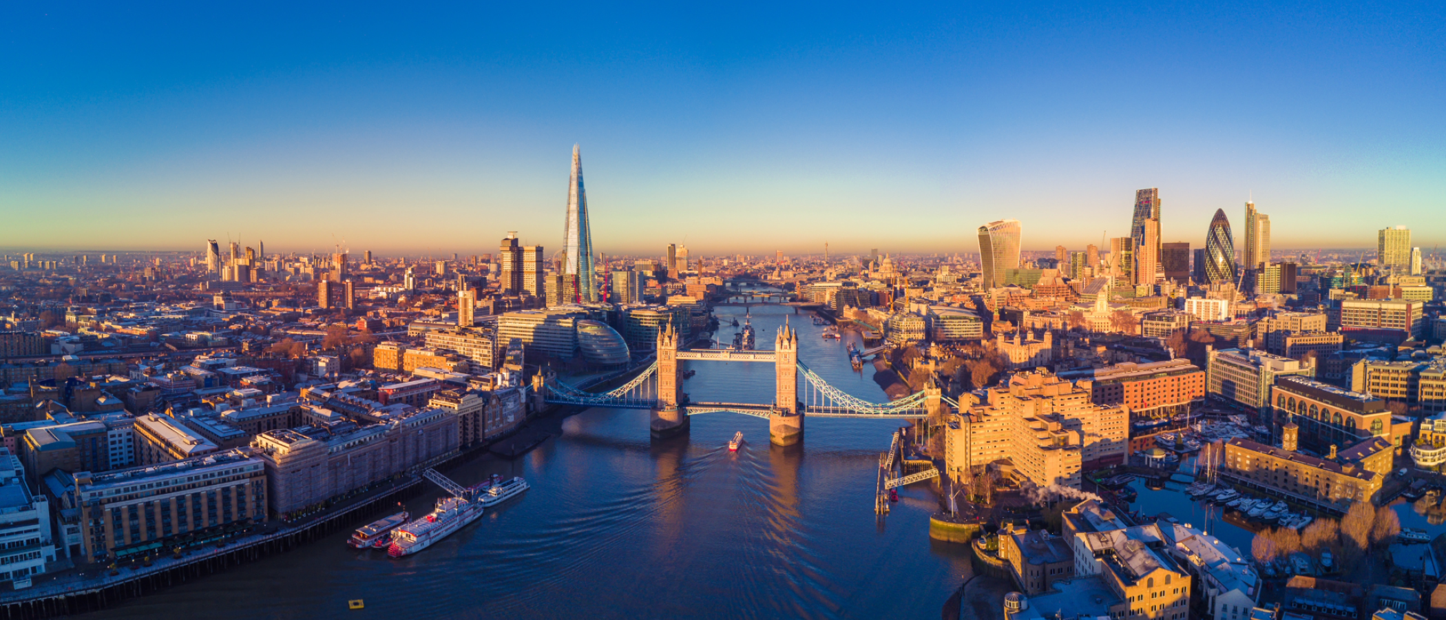 An aerial view of central London showing Tower Bridge spanning the River Thames, with the Shard, the City of London skyline and surrounding buildings visible in clear daylight.