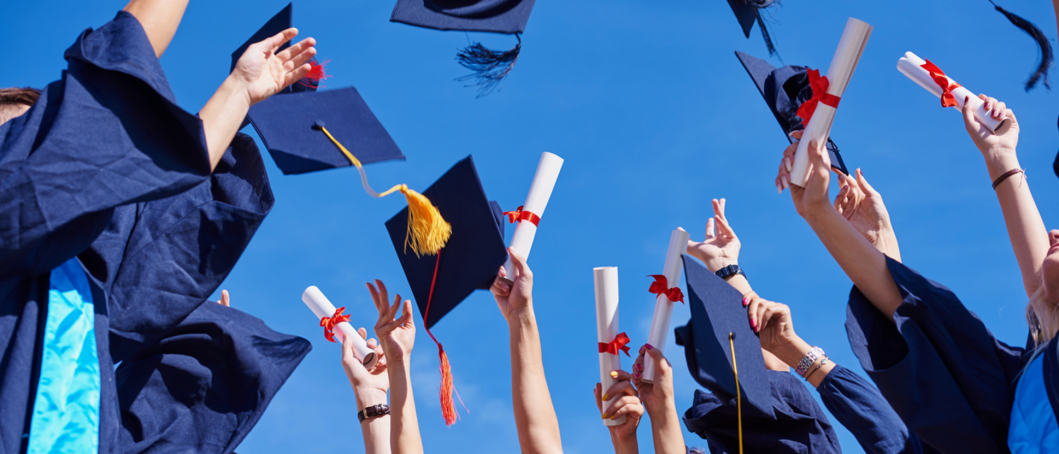 Graduates in caps and gowns celebrate outdoors, throwing graduation caps into the air while holding diploma certificates against a clear blue sky.