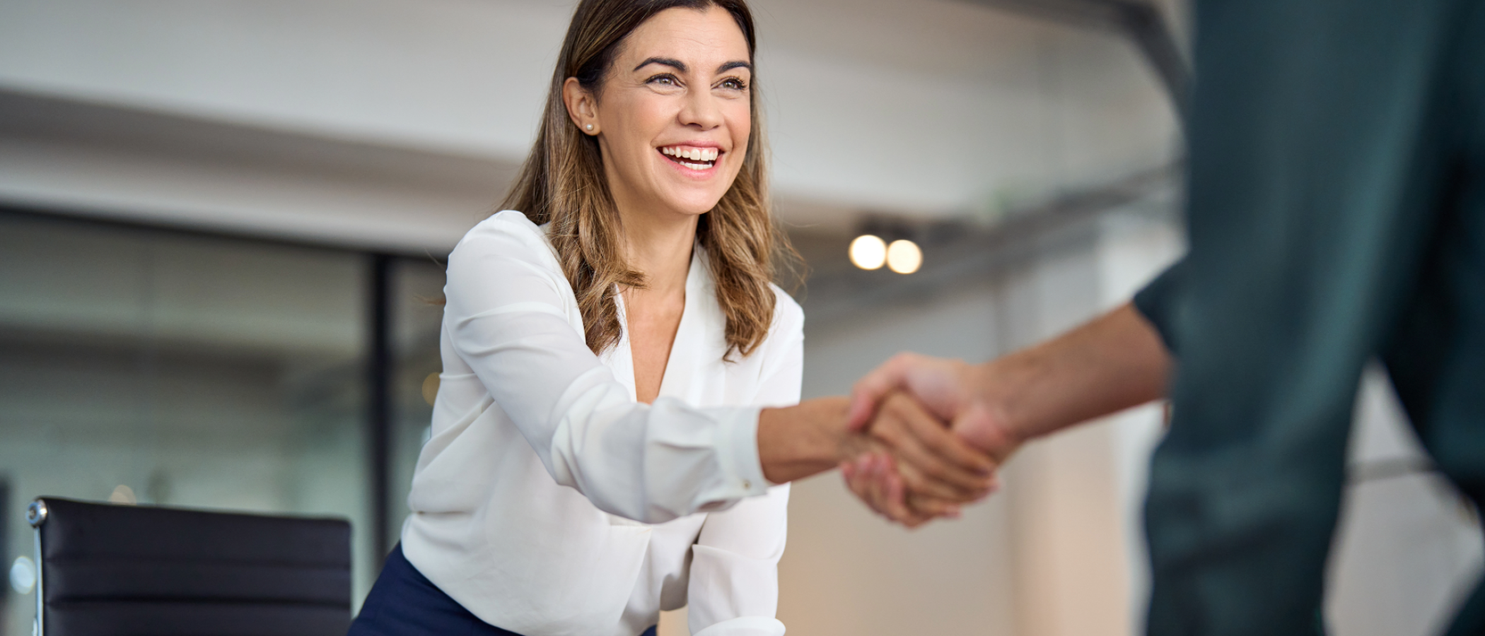 A person leans across a desk to shake hands with another person in a modern office meeting room.