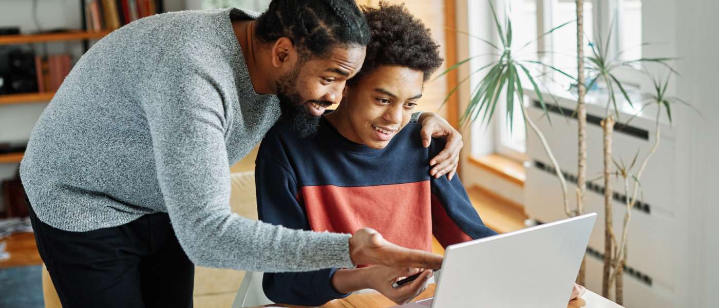 Man and boy looking at a laptop. Boy is sat down at a desk. Man is stood up and hand on boys shoulder