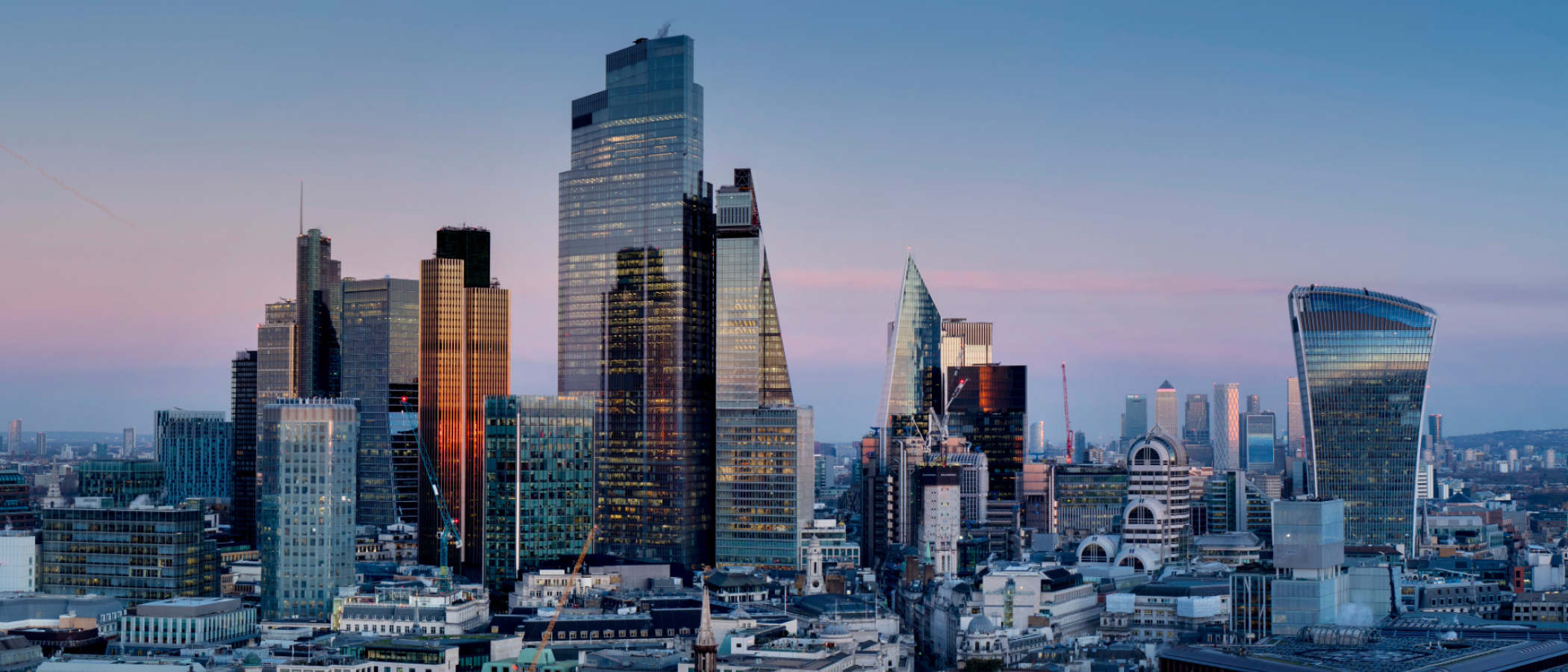 A view of the City of London skyline at dusk, featuring modern skyscrapers including the Walkie Talkie building, surrounded by office towers and historic architecture.