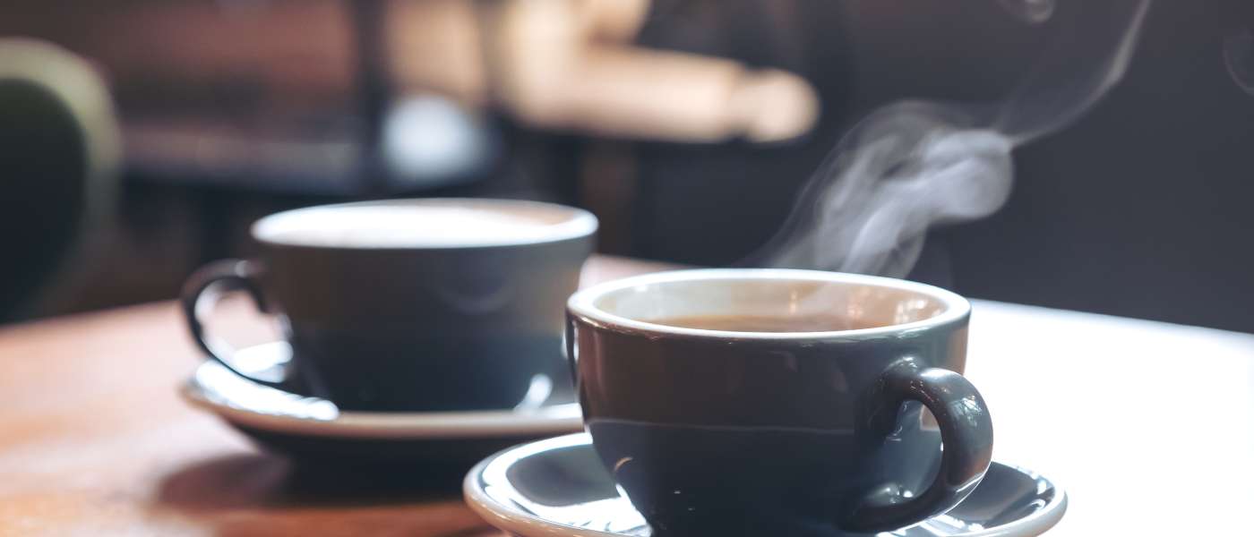 Two black and white coffee mugs and saucers on a brown table. One coffee mug has steam