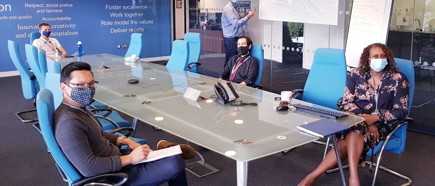 Group of male and female members of staff sitting in meeting room around table wearing covid masks
