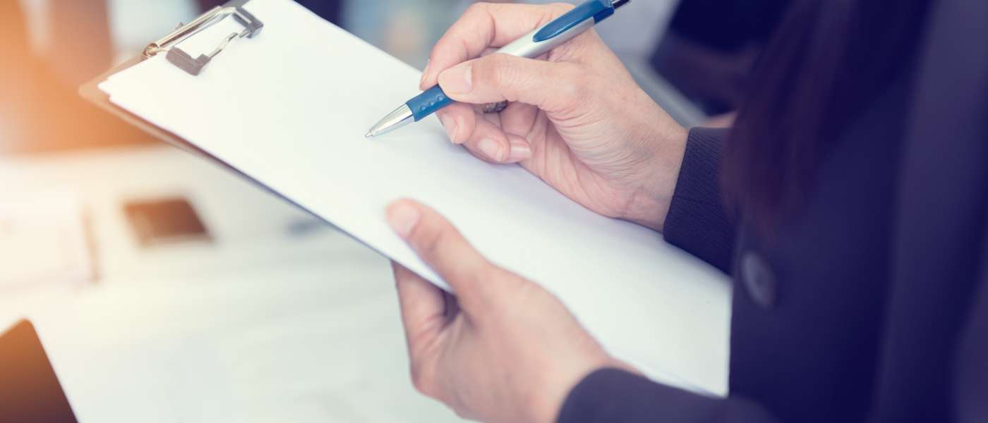 Person wearing a black jacket holding a clipboard and writing on white paper, with a silver and blue pen
