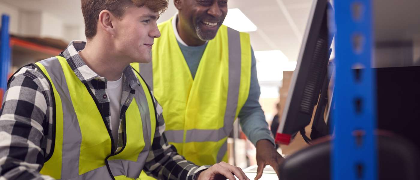 1 younger male and 1 older male working in modern warehouse on computer wearing hi-vis vest