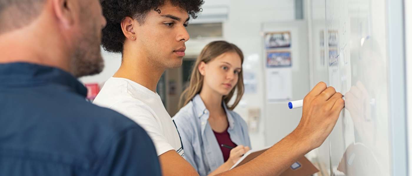 two males and one female are standing in front of a white board. male one is wearing a blue shirt. male two is wearing a white shirt and is writing with a blue marker on the board. female one in a red and blue top is writing in a notebook
