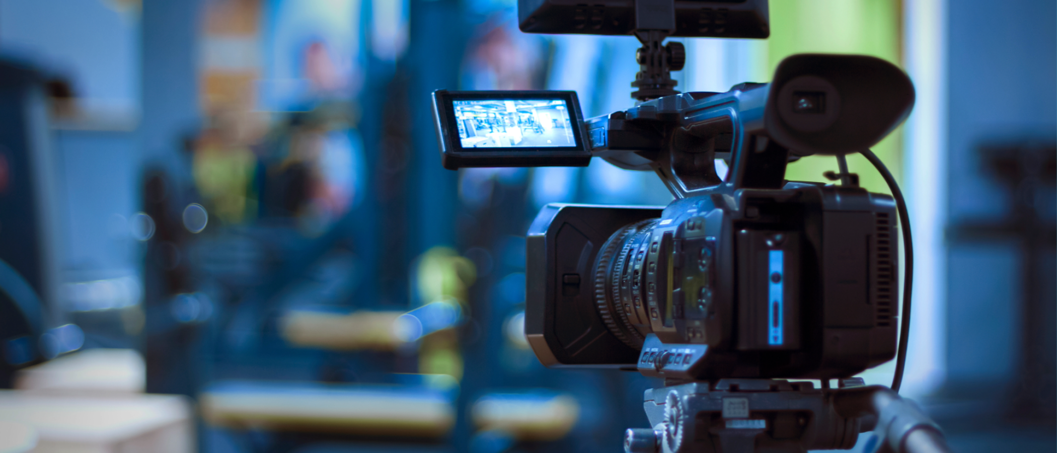 A professional video camera mounted on a tripod records in an indoor studio or production setting, with the camera screen displaying a live view of the scene.