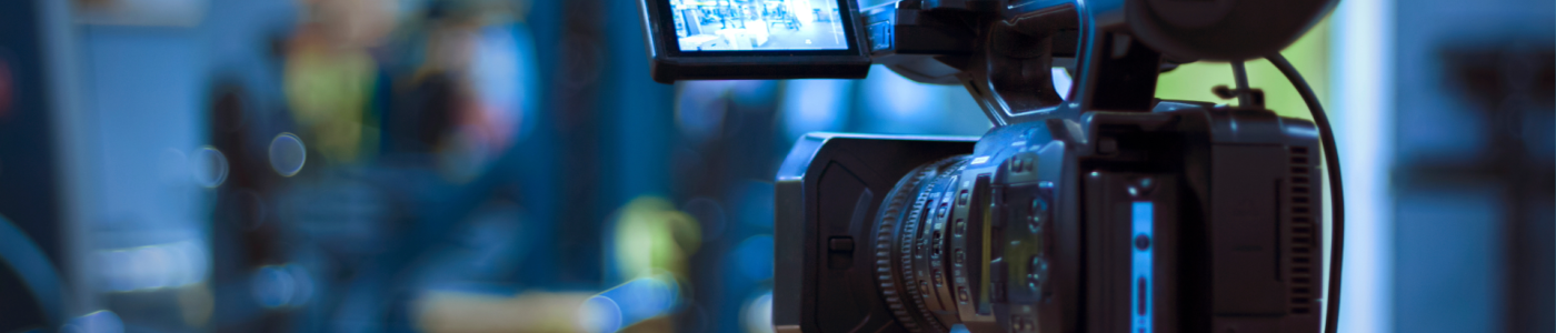 A professional video camera mounted on a tripod records in an indoor studio or production setting, with the camera screen displaying a live view of the scene.