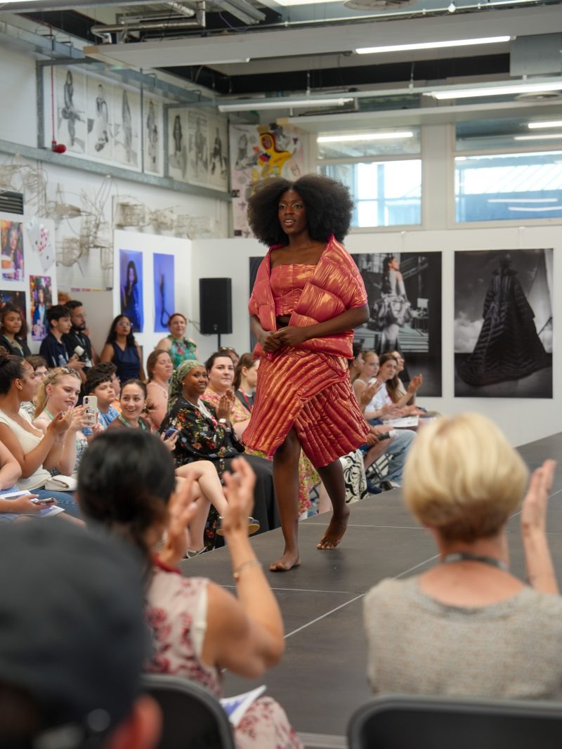 Model in a coral pink outfit walking the runway at a fashion show with an audience seated along the catwalk in an art gallery setting