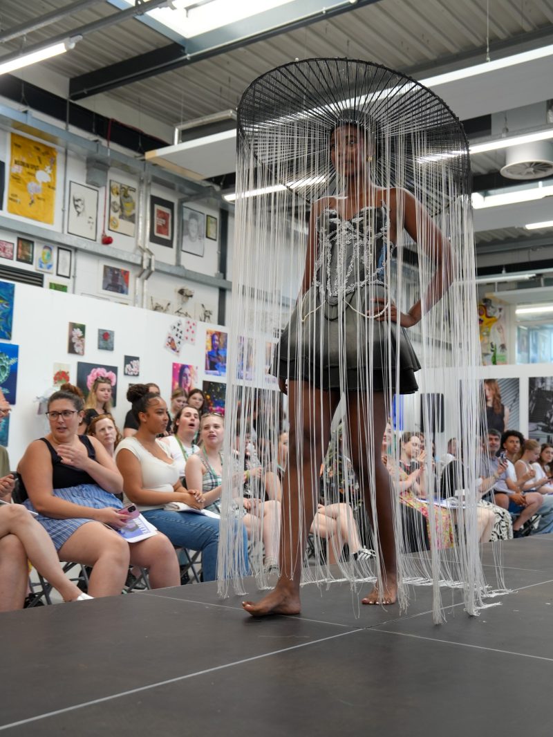 Model wearing a sculptural black and silver outfit with a dramatic fringed circular headpiece on a fashion show runway