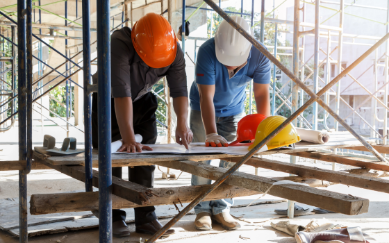 Two construction workers wearing hard hats review building plans on a workbench surrounded by scaffolding at an active construction site.  Provide your feedback on BizChat