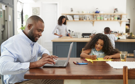 Family in their kitchen man working on laptop at table with girl next to him drawing