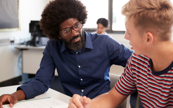 A tutor provides one-to-one guidance to a student at a classroom desk, reviewing written work and offering academic support in a learning environment.