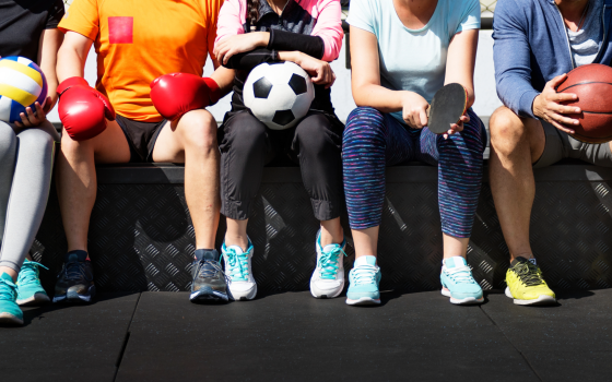 A group of people sit side by side holding different sports equipment, including a volleyball, football, basketball, table tennis bat and boxing gloves, representing participation in a range of sports and physical activities.