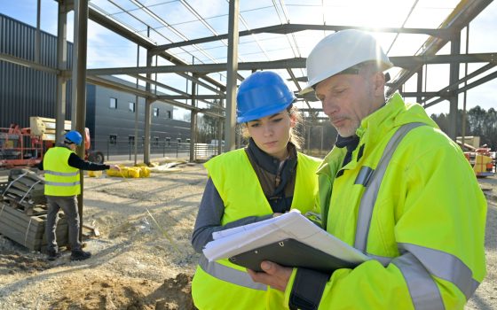 Two construction workers in high visibility clothing and hard hats review building plans at a construction site with another worker measuring in the background