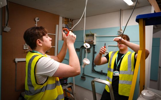 Two young workers in high visibility vests use hand tools to work on electrical wiring inside a training or construction workspace with exposed cables and electrical equipment around them