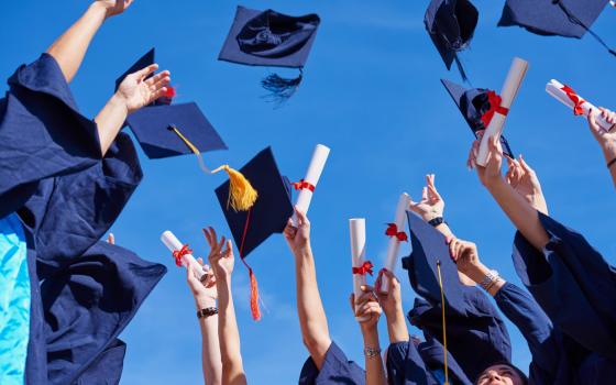 Graduates in caps and gowns celebrate outdoors, throwing graduation caps into the air while holding diploma certificates against a clear blue sky.