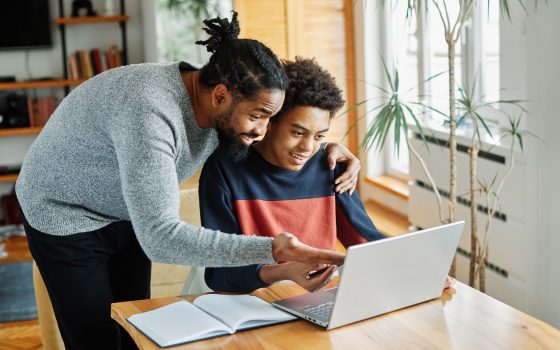 Man and boy looking at a laptop. Boy is sat down at a desk. Man is stood up and hand on boys shoulder