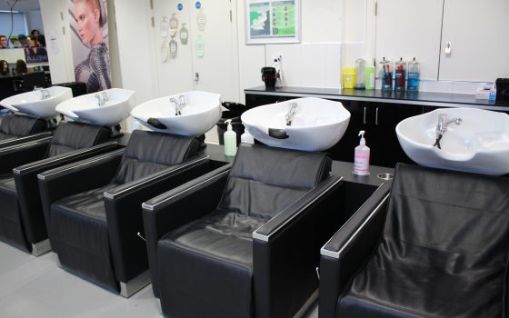 Five black hair salon chairs with attached white sinks. Hair products in the back on a storage unit. Canvas image with a model on the wall and hair mannequins in the far back