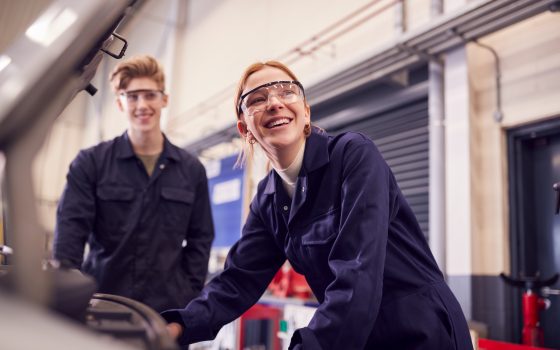 Male And Female students looking at car engine wearing protective overalls and eyewear