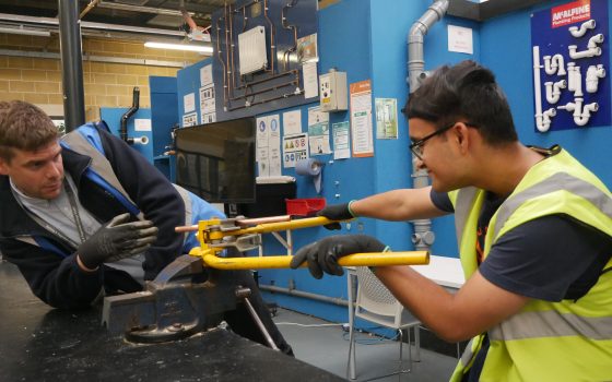 a tutor and student wearing hi vis jackets and gloves in a construction workshop. both are working on construction materials and equipment, helping each other