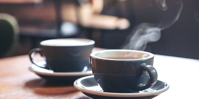 Two black and white coffee mugs and saucers on a brown table. One coffee mug has steam